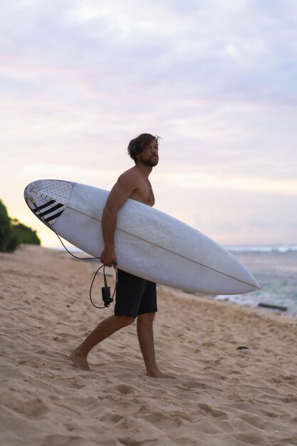 海の波を背景にサーフボードを腕に抱えて日の出のビーチを歩くのに忙しい男性サーファーの風景画像。海の上の若いハンサムな男性サーファー