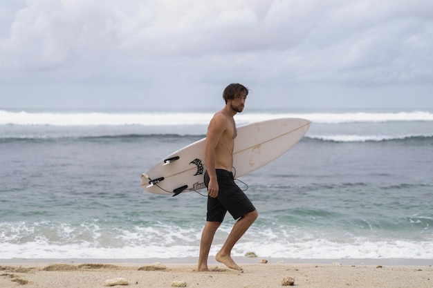 Landscape image of male surfer busy walking on the beach at sunrise while carrying his surfboard under his arm with the ocean waves breaking in the background. Young handsome male surfer on the ocean