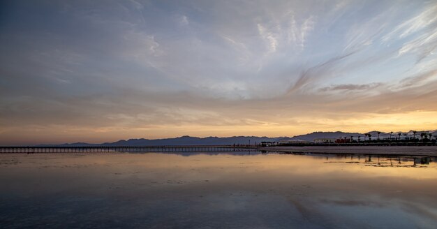 A landscape of the sea with a long pier and mountains on the horizon.