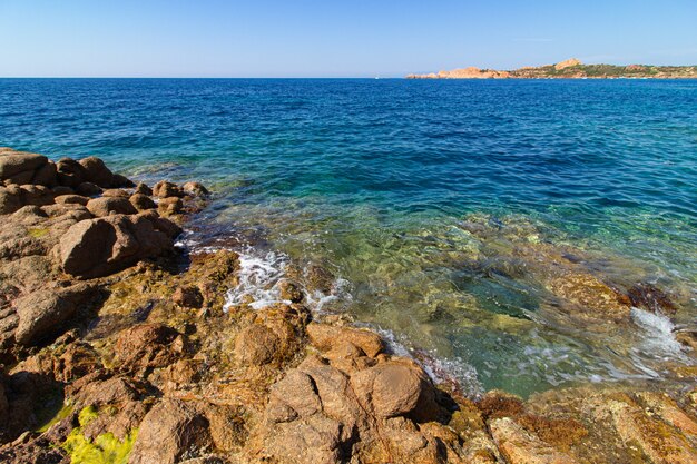 Landscape shot of big rocks, green hills in a blue ocean with a clear blue sky