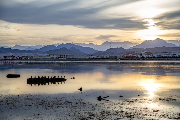 The landscape sky is reflected in the sea in the setting light. City coastline with mountains on the horizon.