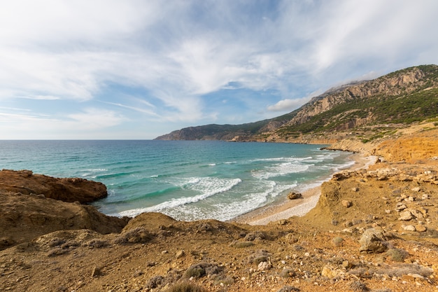 Karpathos 그리스에서 푸른 흐린 하늘 아래 녹지로 둘러싸인 돌 해안의 풍경