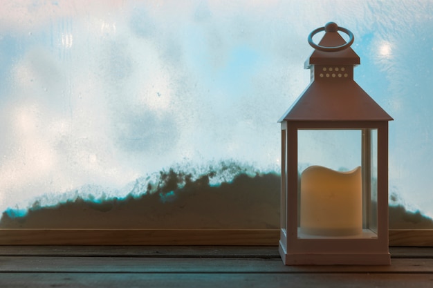 Lantern with candle on wood board near bank of snow through window