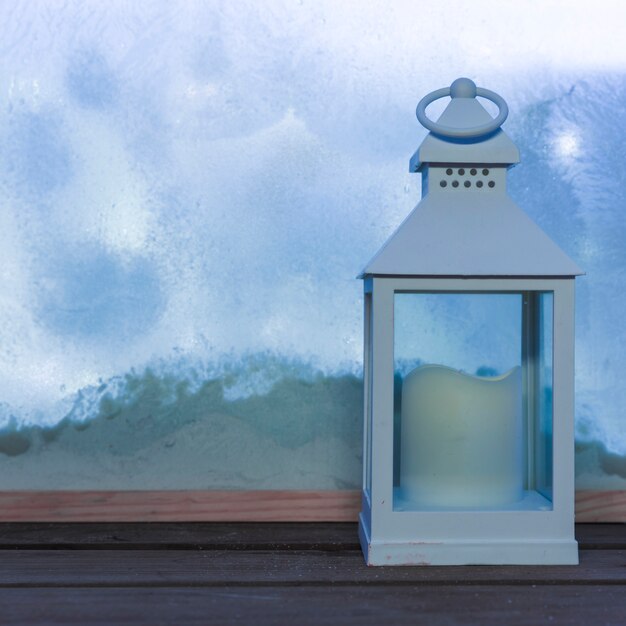 Lantern with candle on wood table near bank of snow through window