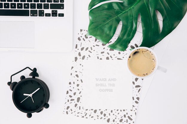 Laptop; alarm clock; monster leaf; coffee cup; adhesive notes with message and paper on white desk