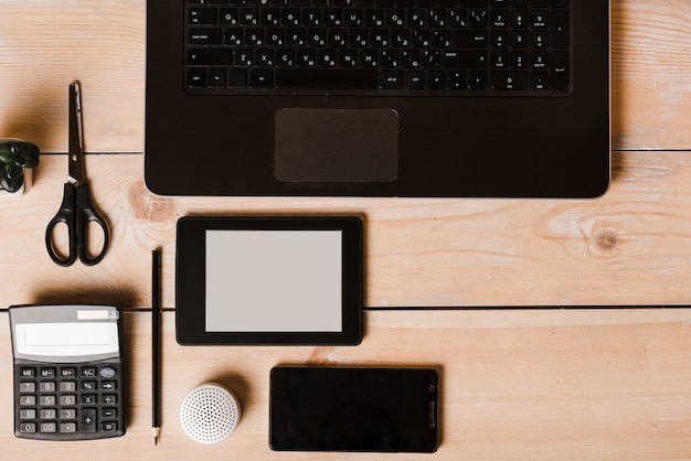 Laptop; calculator; pencil; scissor; cell phone; wireless speaker and ebook reader on wooden desk