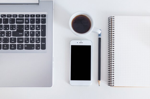 Laptop, cellphone, coffee, pencil and spiral notebook over the white desk