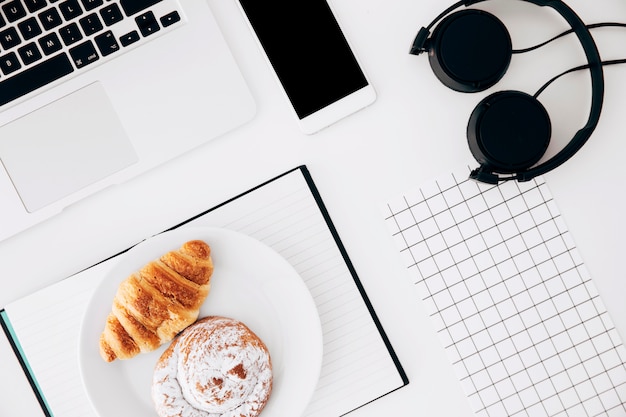 Laptop; cellphone; headphone; square grid paper; baked croissant and buns on diary over white background