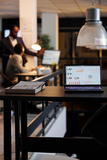 Бесплатное фото Портативный компьютер с финансовыми графиками на экране, стоя на столе в стартовом офисе, коллеги обсуждают стратегию компании на заднем плане. сотрудники, работающие допоздна в стартап-офисе