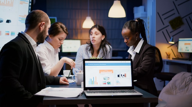 Laptop computer with financial graphs presentation on monitor standing on conference table in meeting room late at night