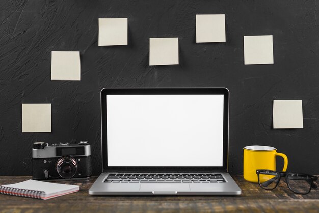 Laptop; cup; spectacles; spiral notepad and camera in front of adhesive notes stucked on blackboard