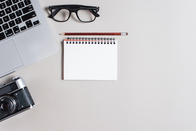 Laptop; eyeglasses; camera; pencil and spiral notepad on gray backdrop