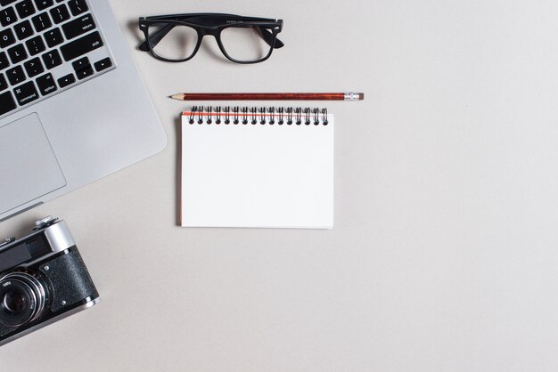 Laptop; eyeglasses; camera; pencil and spiral notepad on gray backdrop