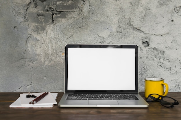 Laptop; spectacles; cup; pencil; spiral notepad and bulldog clip on wooden desk