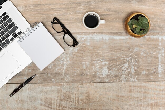 Laptop; spiral notepad; eye glasses; black tea and plant on wooden table