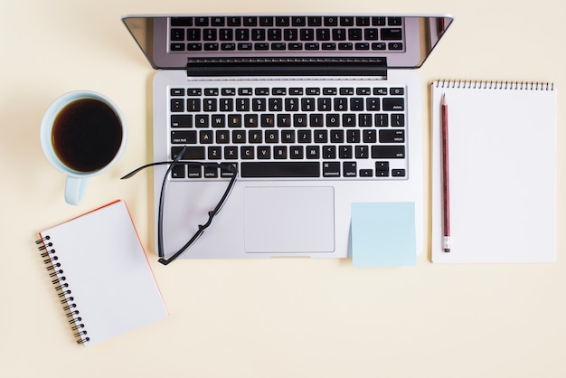 Laptop; tea cup; spiral notepad; eyeglasses and laptop on beige backdrop