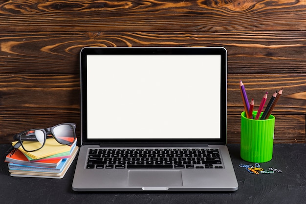 Laptop with blank white screen; books; eyeglasses; pencil holders and paperclip on wooden desk