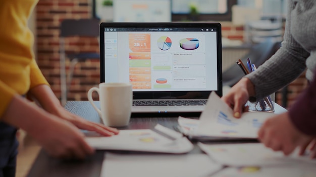 Laptop with financial diagrams and graphs on display in startup company office. Business people using documents with sales reports and analysis, working on partnership growth. Close up