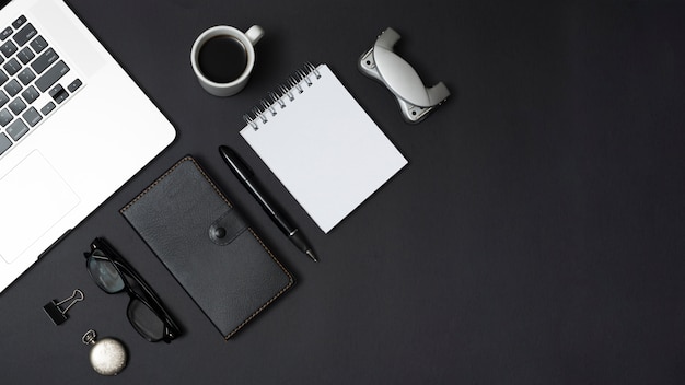 Ноутбук с канцелярскими и личными принадлежностями с чашкой чая на черном фоне