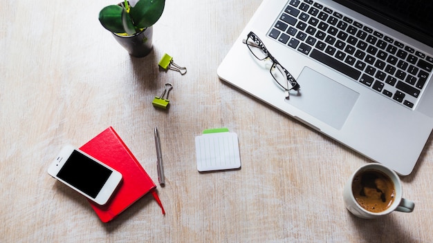 Laptop with spectacles; stationeries; cell phone; coffee cup on table