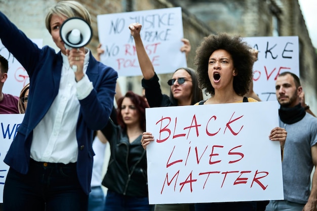 Large group of multiethnic people demonstrating against racism through city streets Focus is on black woman shouting while holding a banner