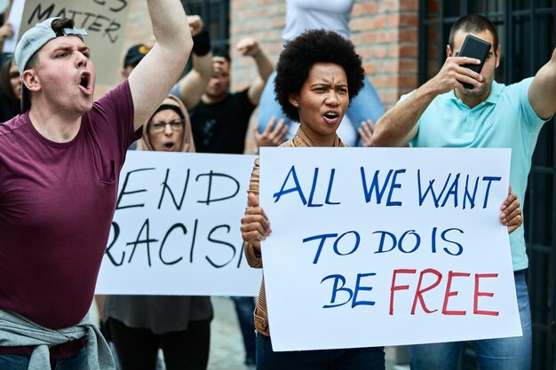 Большая группа людей, протестующих против расизма на улицах. В центре внимания чернокожая женщина с плакатом с надписью о свободе.