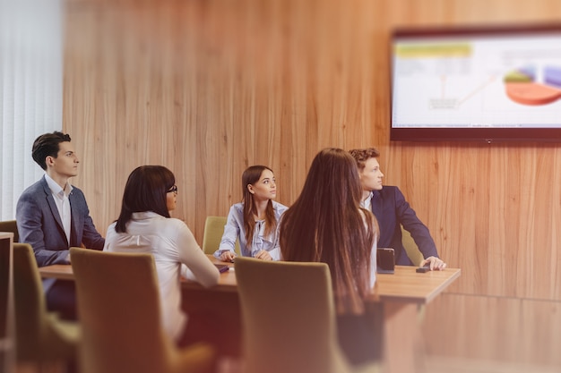 Large team of people is working at one table for laptops, tablets and papers, on the background a large TV set on a wooden wall