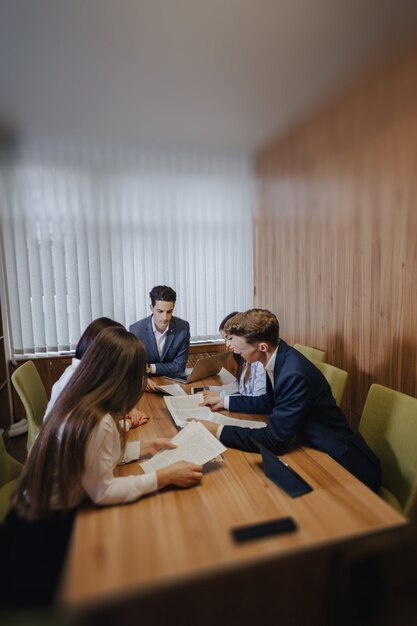 Large team of people is working at one table for laptops, tablets and papers, on the background a large TV set on a wooden wall