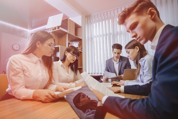 Large team of people is working at one table for laptops, tablets and papers,  a large TV set on a wooden wall