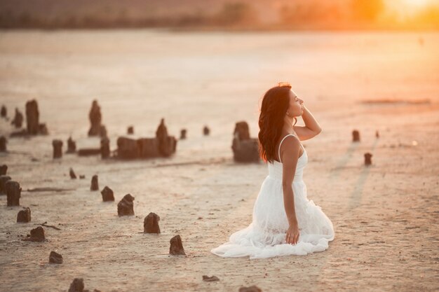 석양에 모래에 무릎에 앉아있는 갈색 머리 소녀의 측면보기