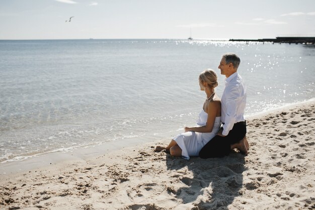 Lateral view of couple which is sitting on the sandy beach near the sea and looking on the breathtaking landscape