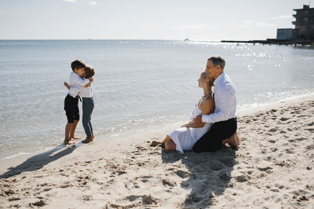 Lateral view of couple which is sitting on the sandy beach near the sea and looking on the two little sons who are hugging