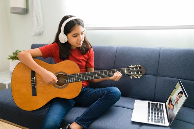 彼女のオンライン音楽レッスンを聞いているヘッドフォンを持つラテン系の女の子。アコースティックギターを弾き、和音を学ぶ芸術的な子供