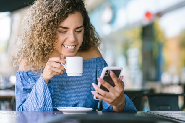 コーヒーショップでコーヒーを飲みながら携帯電話を使用しているラテン女性。アーバンコンセプト。