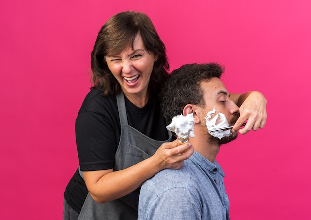 Ridendo adulto femmina caucasica barbiere in uniforme che tiene pennello da barba con schiuma e barba da barba di giovane uomo con rasoio isolato su sfondo rosa con spazio di copia