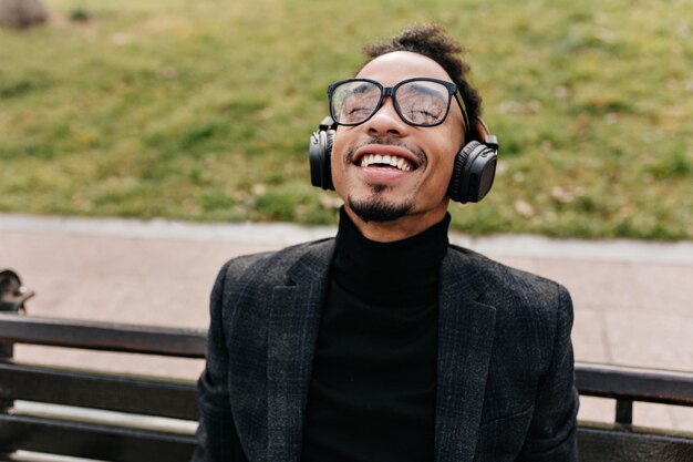 Laughing african man posing on wooden bench with green lawn . Happy black guy in glasses listening music with eyes closed and smiling.