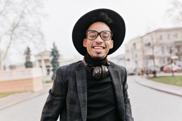 Laughing african man with trendy curly hairstyle wearing hat. Outdoor photo of male model with dark skin having fun while exploring town.