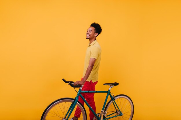 Laughing african young man in stylish pants posing with bicycle Studio shot of goodlooking male model standing on yellow background with smile