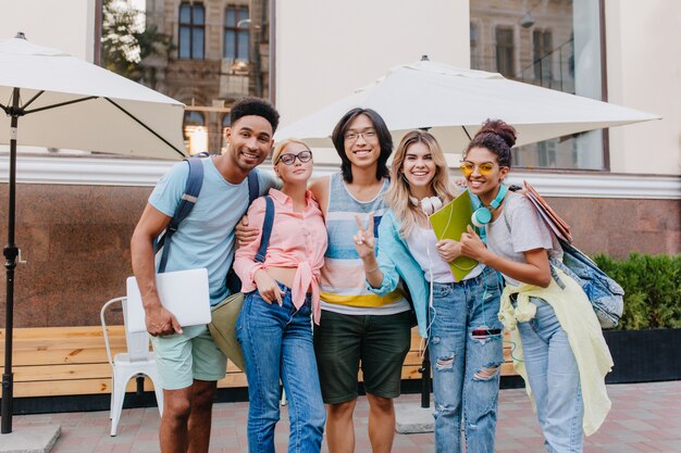 Laughing asian boy in glasses and shorts embracing charming blonde girls in front of outdoor cafe. Joyful students came to open-air restaurant to celebrate end of exams