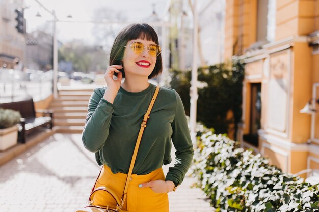 トレンディな春の服装で魅力的な女の子を笑って屋外で冷やします。幸せな白人女性の写真はサングラスと黄色のハンドバッグを身に着けています。