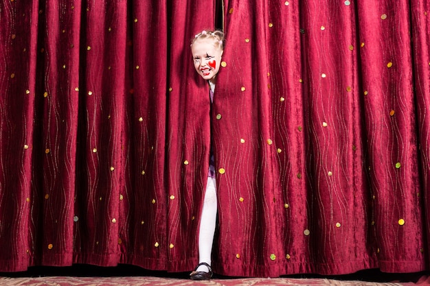 Смеющаяся привлекательная молодая блондинка с красочным макияжем выходит на сцену во время спектакля или пантомимы, переступая через закрытые шторы