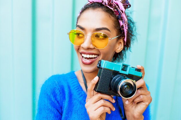 Laughing black girl with cute hairstyle holding retro camera and looking at camera.