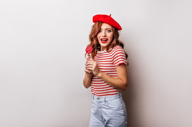 Laughing blissful girl holding sweet candy. Indoor photo of carefree caucasian woman in beret posing with lollipop.