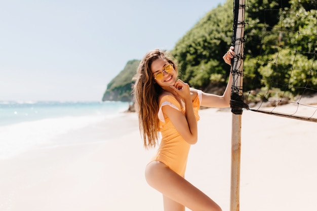 Смеющаяся блаженная девушка в солнечных очках танцует возле волейбольной площадки на курорте. Красивая кавказская женская модель в купальнике дурачится на экзотическом пляже.