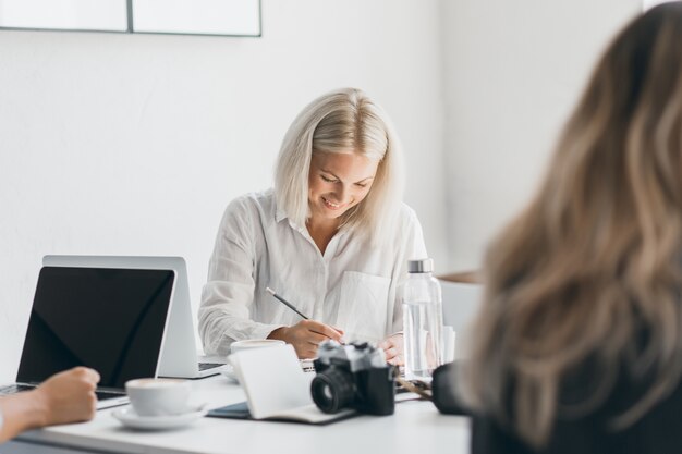 Laughing blonde woman in white shirt looking down while writing something. Indoor portrait of busy female freelance specialist posing at workplace with laptop and camera.
