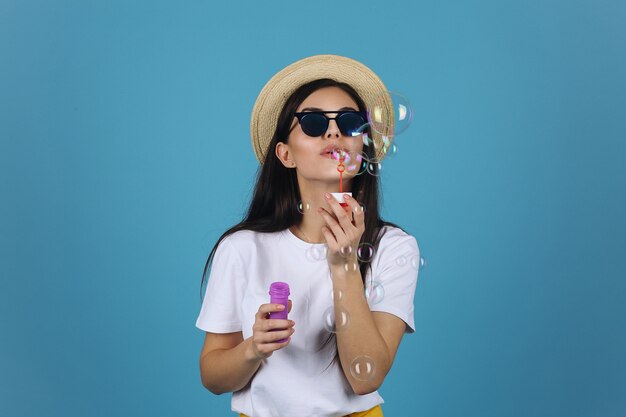 Laughing brunette girl in summer hat and sunglasses has fun with soap balloons