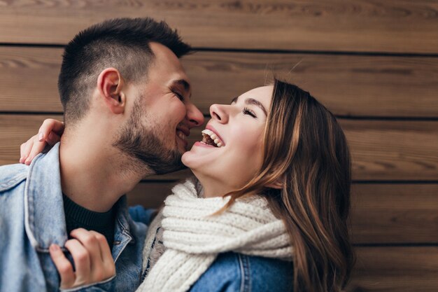 Laughing brunette man expressing love while posing with girlfriend. Blissful caucasian couple kissing on wooden wall.