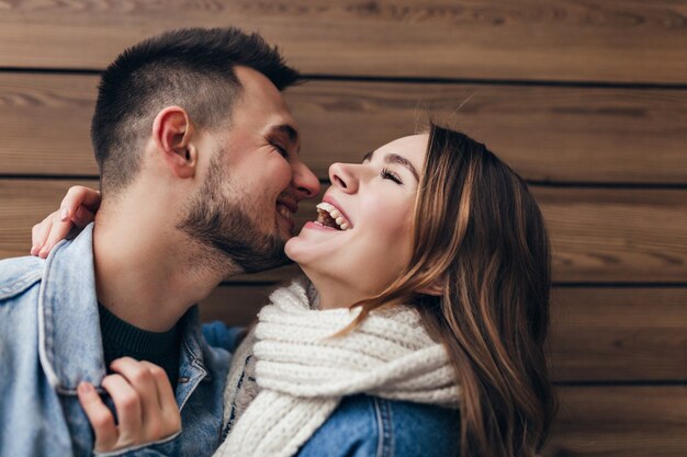 ガールフレンドとポーズしながら愛を表現するブルネットの男を笑う。木製の壁にキスする至福の白人カップル。