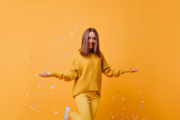 Бесплатное фото Смеющаяся брюнетка в желтых штанах выражает счастье и выбрасывает конфетти. фотография в помещении привлекательной хорошо одетой девушки, танцующей на яркой стене.
