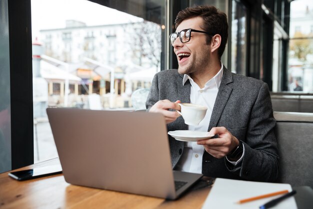 Laughing businessman in eyeglasses sitting by the table in cafe with laptop computer and cup of coffee while looking at window